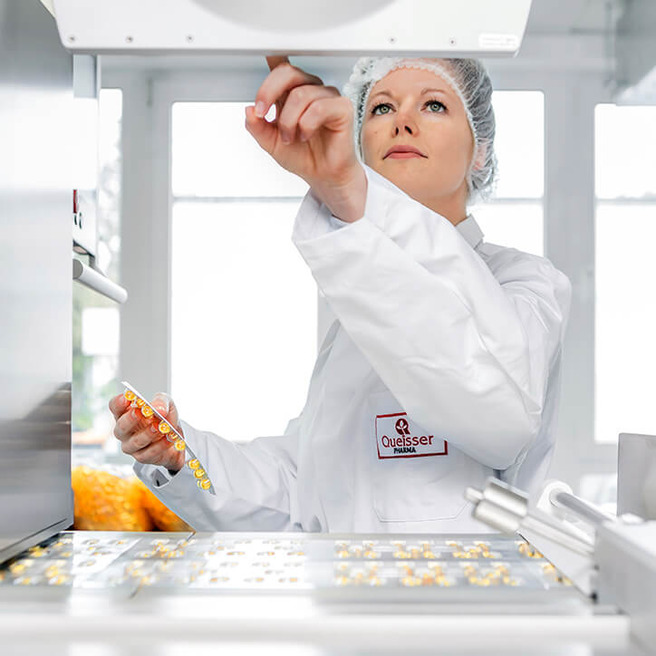 Ženska v beli delovni obleki s pokrivalom na glavi se osredotoča na stroj v laboratoriju. Z eno roko drži paket, medtem ko drugo dviga. Ozadje prikazuje svetlo okolje s okni in elemente opreme.