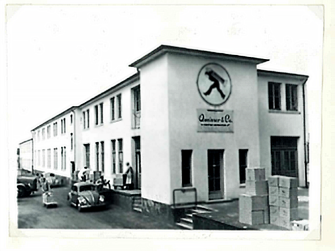 Zgodovinska črno-bela fotografija stavbe s podjetjem "Quinver & Co" na fasadi. Pred stavbo so parkirani starinski avtomobili, ljudje pa opravljajo različne dejavnosti ob skladiščenju škatel.