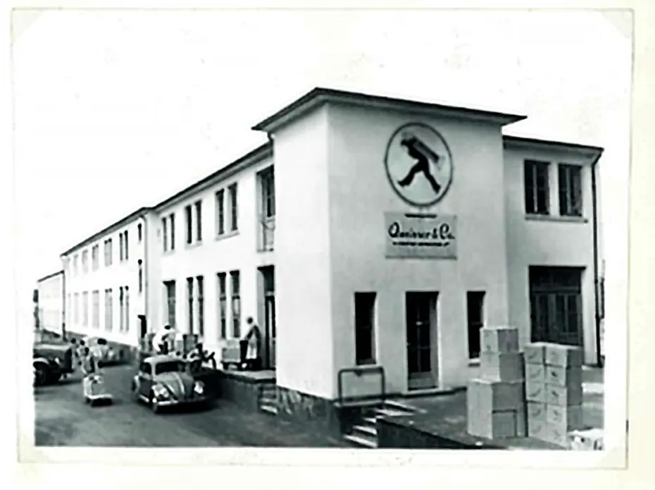 Zgodovinska črno-bela fotografija stavbe s podjetjem "Quinver & Co" na fasadi. Pred stavbo so parkirani starinski avtomobili, ljudje pa opravljajo različne dejavnosti ob skladiščenju škatel.