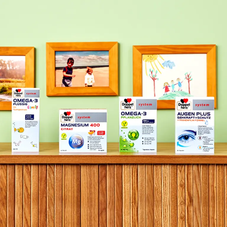 Na polici so štiri škatle prehranskih dopolnil: OMEGA-3 FLUID, MAGNESIUM 400 CITRAT, OMEGA-3 RASTLINSKI in AUGEN PLUS. V ozadju so otroške risbe in fotografije družinskih članov. Stena je v nežno zeleni barvi.