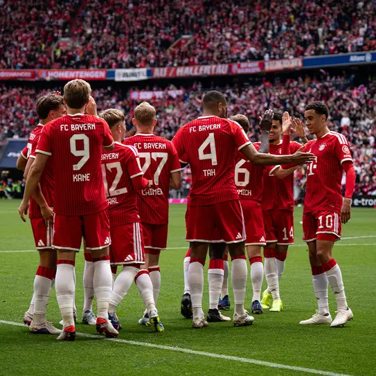 Ekipa FC Bayern se zbere na igrišču, se pozdravlja in razpravlja. Igralci nosijo rdeče drese s številkami ter se gibljejo okoli, medtem ko je v ozadju navijaška množica.