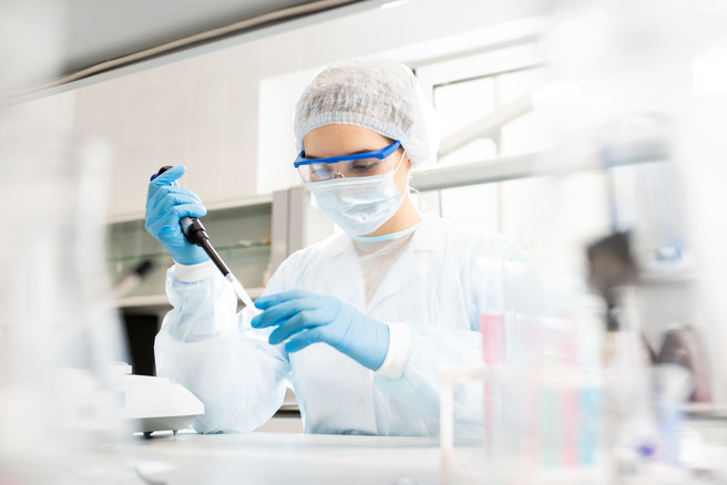 Znanstvenik v laboratorijski opravi z zaščito za obraz in rokavicami uporablja pipeto za merjenje tekočine v laboratoriju. V ozadju so različni laboratorijski pripomočki, ki podpirajo raziskovalno delo.
