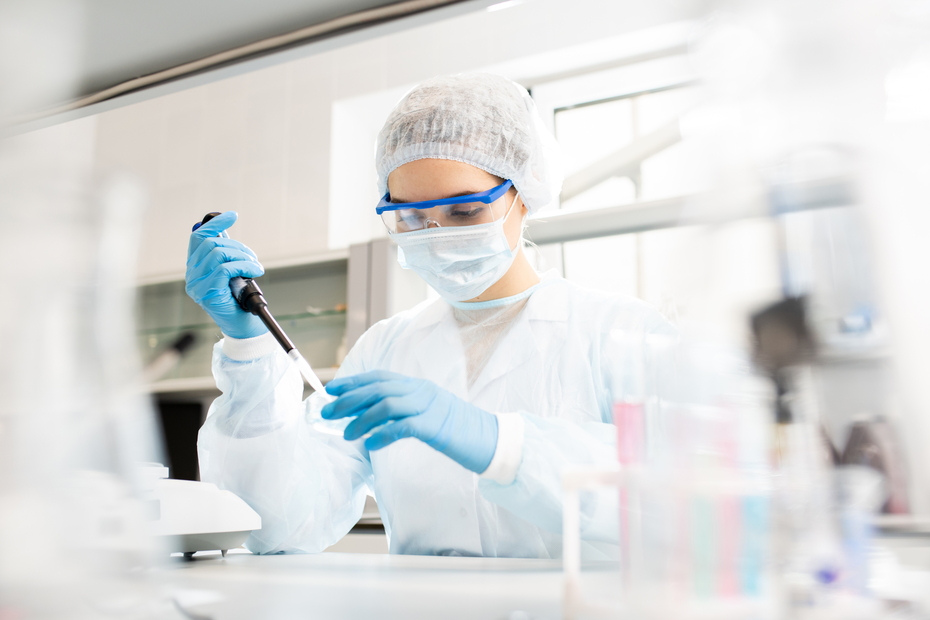 Znanstvenik v laboratorijski opravi z zaščito za obraz in rokavicami uporablja pipeto za merjenje tekočine v laboratoriju. V ozadju so različni laboratorijski pripomočki, ki podpirajo raziskovalno delo.