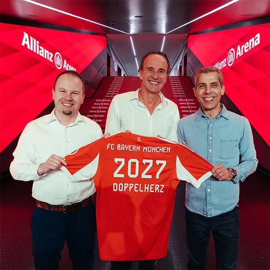 Na sliki so trije moški, ki se nahajajo v Allianz Areni. Držijo dres FC Bayern Munchen, ki je oranžne barve in ima napis "2027 DOPPELHERZ". V ozadju so svetleče rdeče stene stadiona.