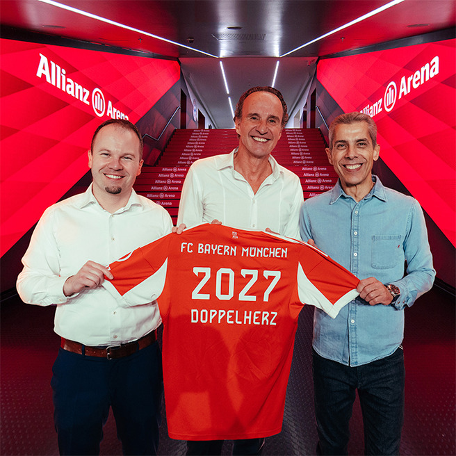 Na sliki so trije moški, ki se nahajajo v Allianz Areni. Držijo dres FC Bayern Munchen, ki je oranžne barve in ima napis "2027 DOPPELHERZ". V ozadju so svetleče rdeče stene stadiona.