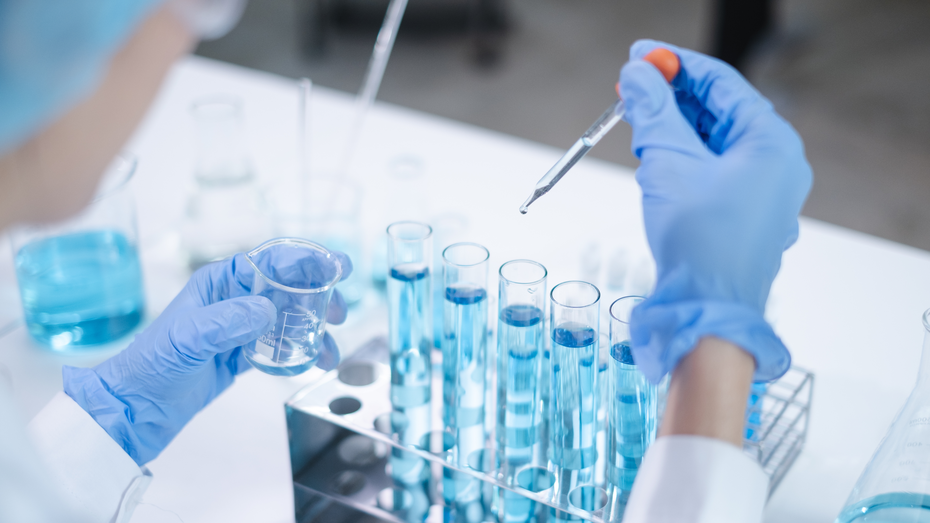 Znanstvenik v laboratoriju drži epruveto z modro tekočino, medtem ko pipetira kapljico iz druge epruvete. Oprema in stekleni posodi so razporejeni na površini mize.