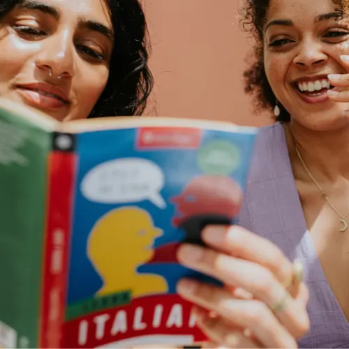 Dve ženski gledata v knjigo z naslovom "Italiano". Ena ženska, ki ima temne lase in nosi nosno nakit, bere, medtem ko druga, z rjavimi kodri, z nasmehom opazuje. Ozadje je rahlo rožnato.