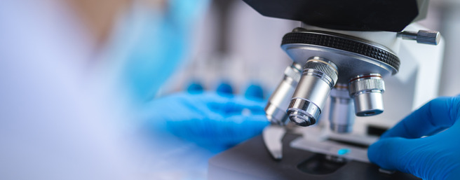 Mikroskop z osebo v zaščitnih rokavicah, ki pripravljajo vzorec za analizo. Oprema je dobro osvetljena, v ozadju so vidni laboratorijski elementi.