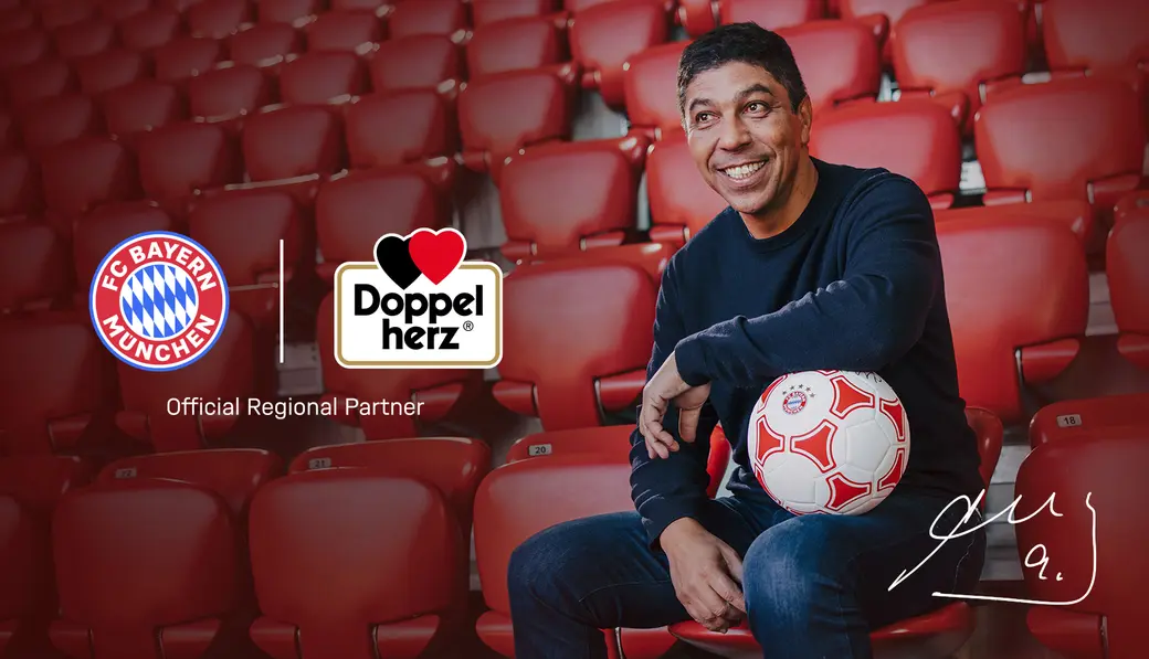 Na sliki je nasmejan moški, ki sedi na sedežih na stadionu. Drži nogometno žogo in je oblečen v črno majico ter modre hlače. V ozadju so logotipi FC Bayern München in partnerja Doppelherz.