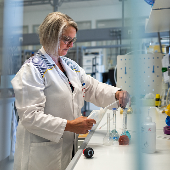 Ženska v laboratorijskem plašču izvaja postopke s steklenimi posodami. Okoli nje so različne laboratorijske naprave in steklenice. Vidi se, da je pozorna na delo, ki ga opravlja.